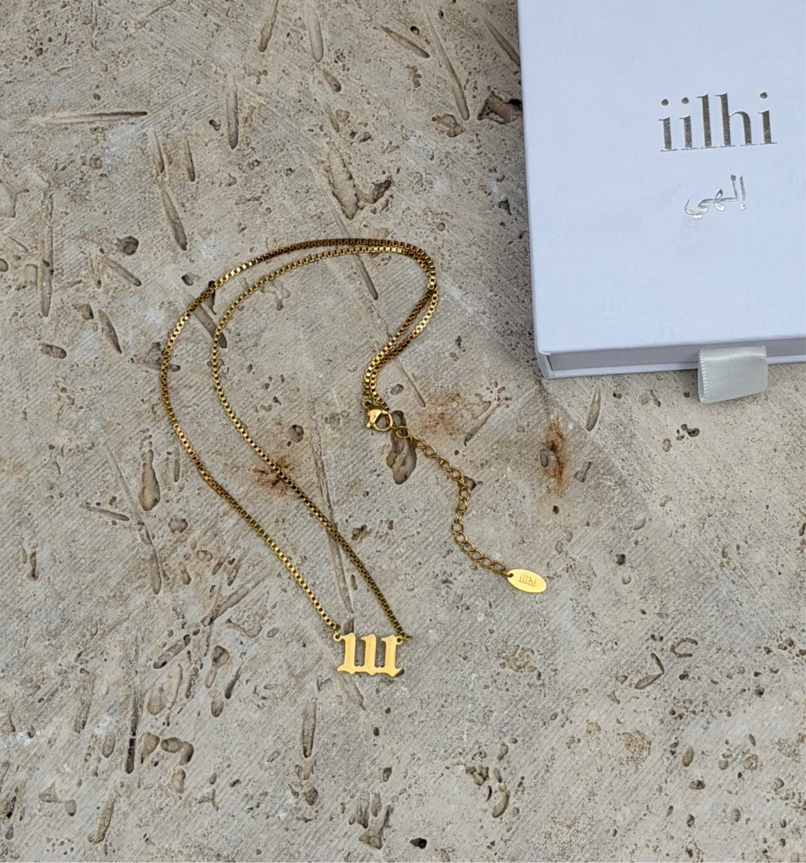 Gold necklace with a '111' pendant on a textured surface next to a box labeled 'iilhi'.