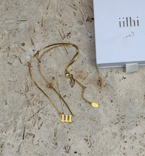 Gold necklace with a '111' pendant on a textured surface next to a box labeled 'iilhi'.