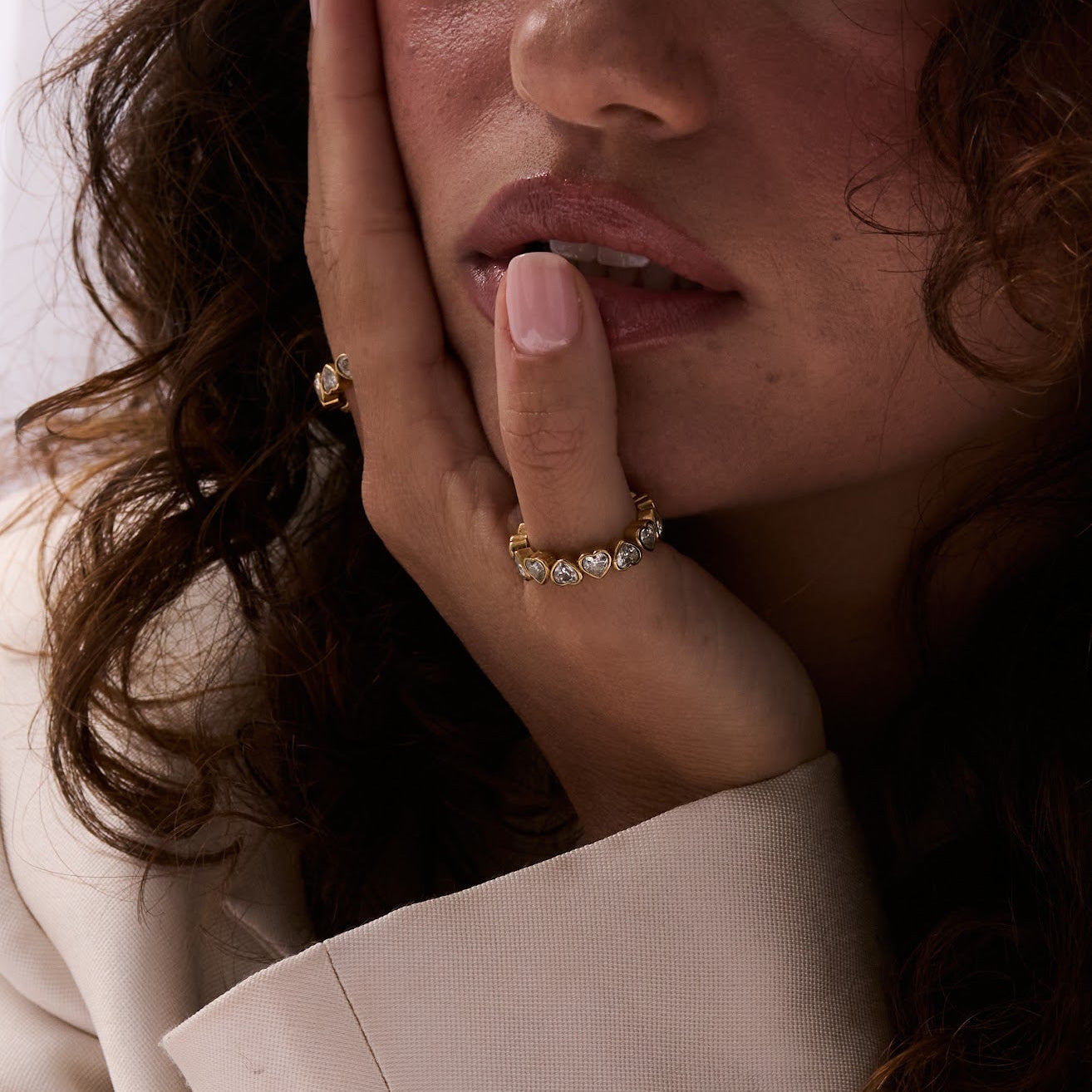 Woman with curly hair wearing a beige blazer, heart ring, close-up of face and hand.